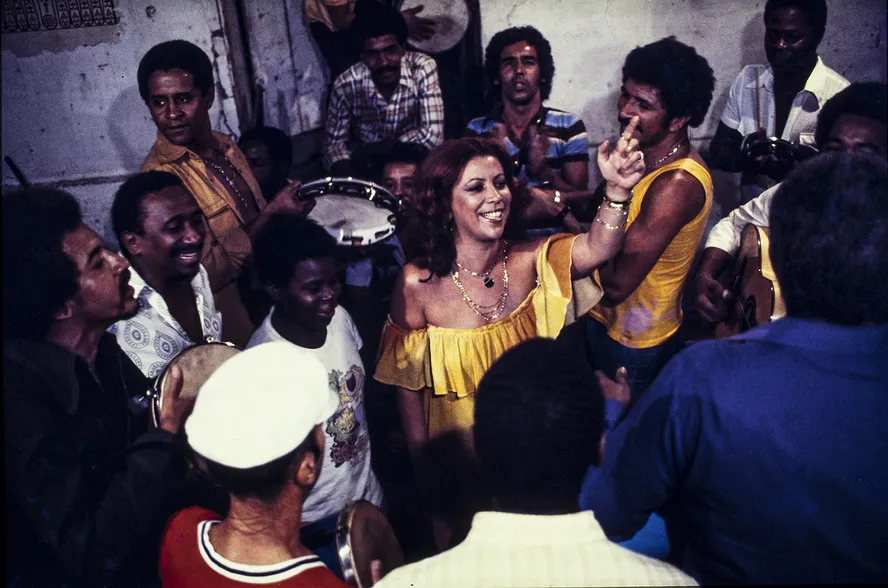 Roda de samba: músicos reunidos cantando e tocando pandeiros, cavaquinho e violão.